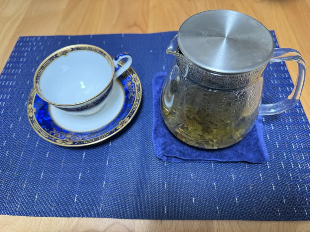青い敷物の上に置いた、茶葉の入ったガラスのティーポットと、青と金の模様のティーカップ